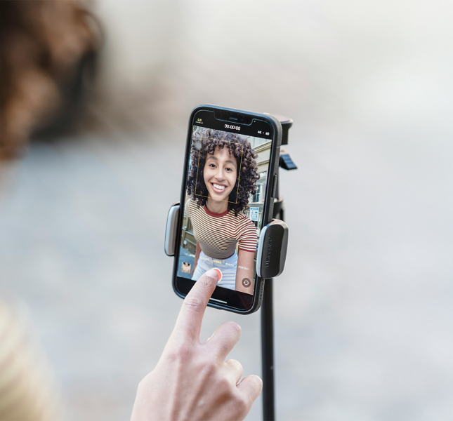 A close-up image of a smartphone mounted on a tripod, capturing a cheerful woman with curly hair wearing a striped shirt during a video recording. A hand is visible in the foreground, about to tap a button on the phone screen.