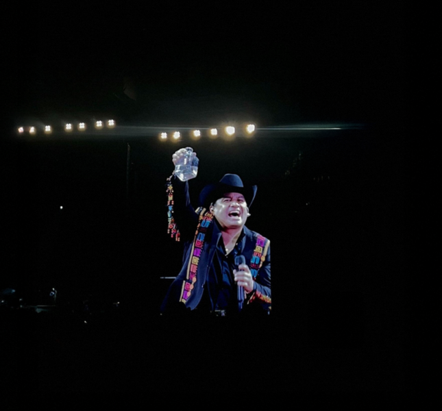 Julión Álvarez, wearing a black cowboy hat and a colorful embroidered jacket, performs on stage at SoFi Stadium. He is smiling, holding a microphone in one hand and raising a bottle in the other. Bright stage lights shine behind him, and the background is mostly dark.