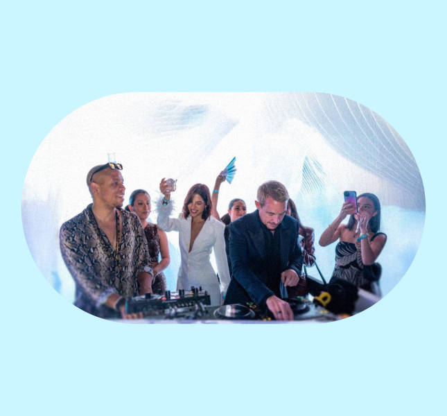 A DJ in a dark suit plays music at a brightly lit event, while another individual in a patterned shirt and sunglasses stands to his left at the DJ booth. Several people are visible behind them, dancing and holding up their phones.