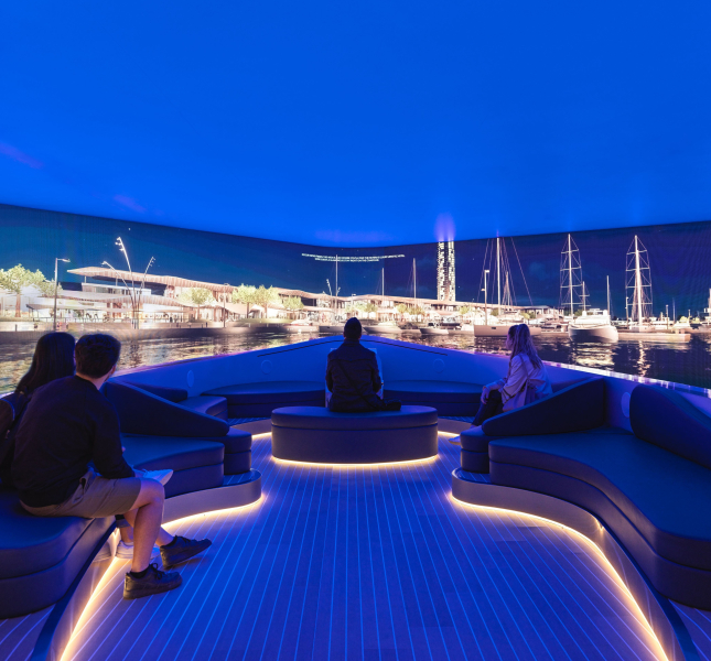 A group of visitors are seated on curved, illuminated benches inside an immersive experience room designed to look like the deck of a yacht. They are watching a panoramic, wrap-around screen displaying a vibrant, photorealistic marina at night.