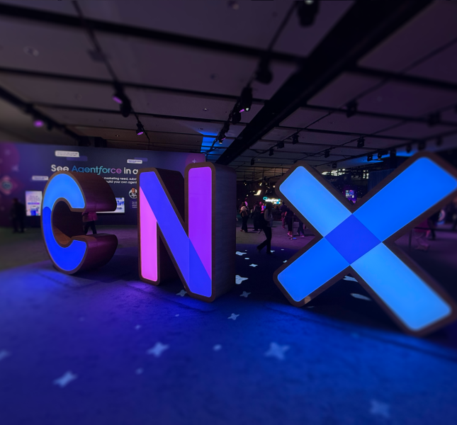 A low-angle, wide shot of large, illuminated, three-dimensional letters spelling "CNX" in a dimly lit event hall. The 'C' glows blue, the 'N' glows purple, and the 'X' glows with a bright blue light. The letters are placed on a dark blue carpet with a subtle pattern, and they cast colorful reflections on the floor. In the blurred background, people can be seen walking around the conference space.