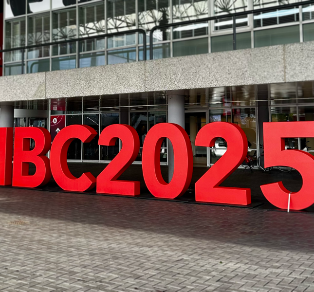 Large, three-dimensional red letters spelling "IBC2025" stand on a brick plaza in front of the entrance to a modern convention center with a glass facade.