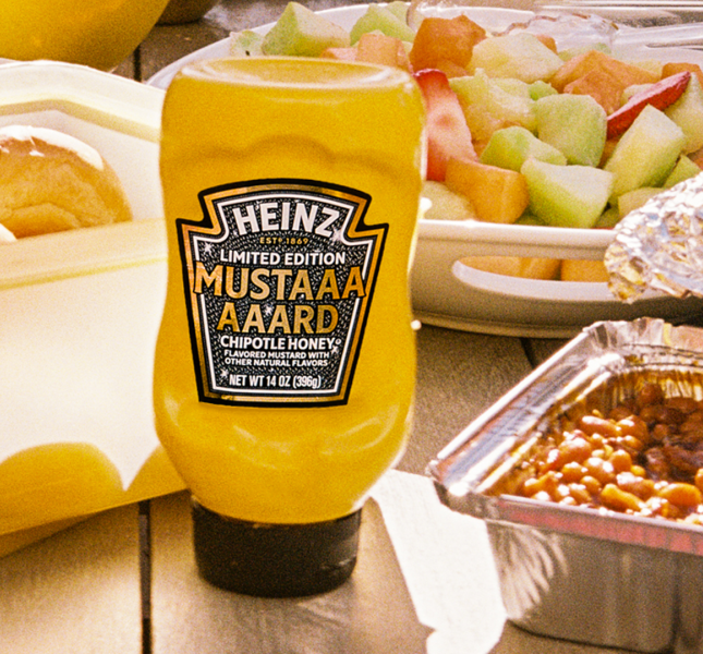Bright yellow Heinz Limited Edition Mustaaaaard Chipotle Honey bottle, prominently displayed on a wooden picnic table next to a fruit salad, baked beans, and fresh buns, highlighting the perfect summer BBQ condiment.