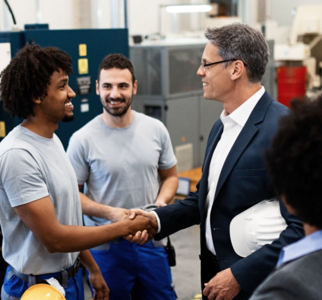 Businessmen shaking hands in a factory. Two factory workers and two people in suits are collaborating.