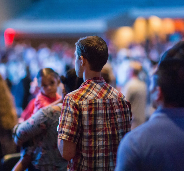 A rear view of a standing crowd in a large, dimly lit indoor venue, focusing on a man in a plaid shirt in the center. To his left, a woman holds a small child who is looking sideways, while the foreground and background contain other attendees blurred out of focus.