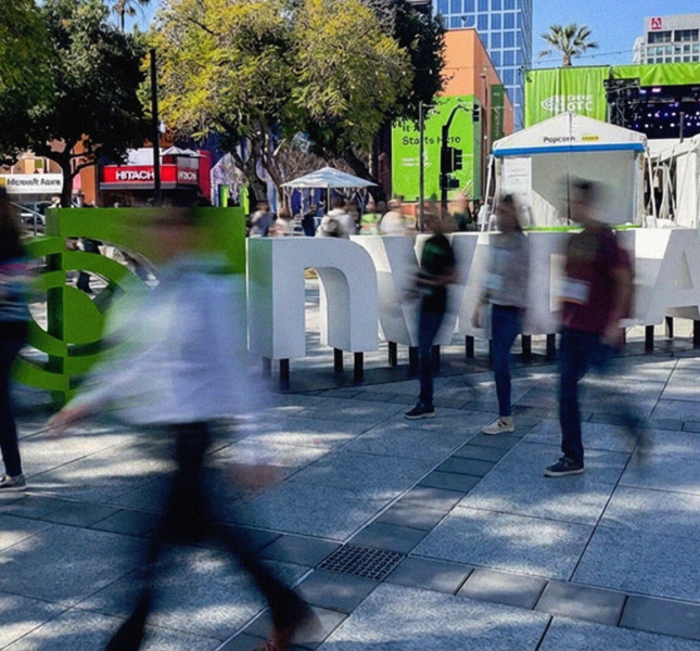A wide-angle, slightly blurred shot of an outdoor plaza at the NVIDIA GTC 2026 conference in San Jose. Large, 3D white letters spelling out "NVIDIA" stand in the center, with the green NVIDIA logo to the left. People are captured in motion, appearing as blurred figures walking across the stone-tiled ground, creating a sense of a busy, electric atmosphere. In the background, there are green banners, white event tents, trees, and city buildings under a clear blue sky.