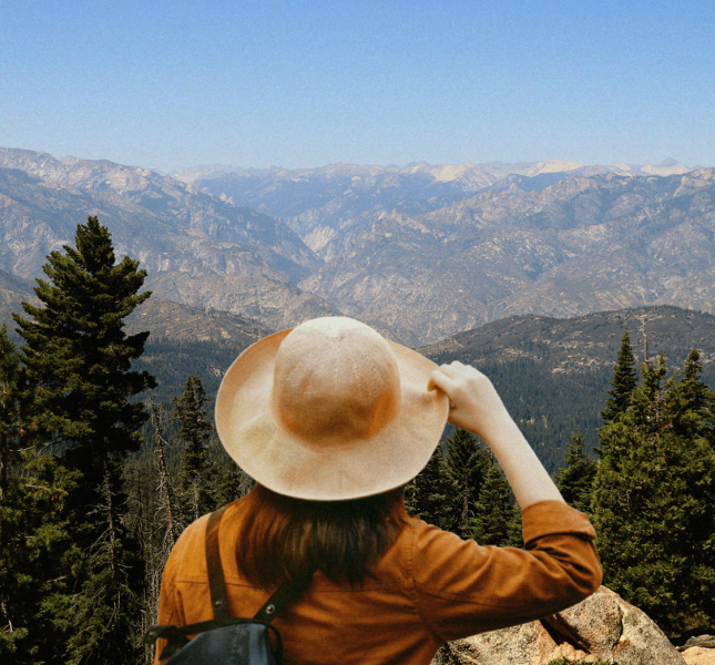 A person seen from behind wearing a wide-brimmed tan hat and an ochre jacket, gazing out at a vast, rugged mountain range and evergreen forest under a clear blue sky.