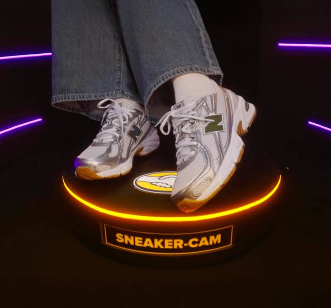 A close-up of a person's feet wearing blue jeans, white socks, and silver and white New Balance sneakers featuring a green "N" logo. They are standing on a small, circular black platform that has a glowing orange ring light around its top edge. The front of the platform displays a yellow label reading "SNEAKER-CAM".