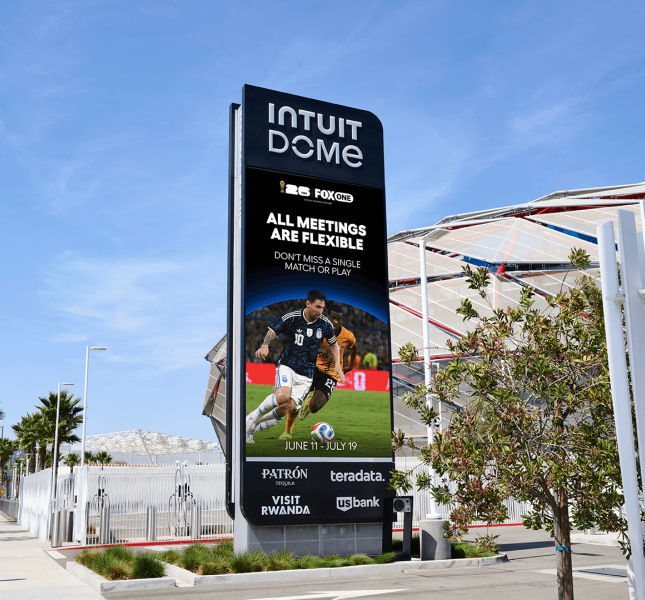 A tall digital billboard outside the Intuit Dome displaying a FIFA World Cup advertisement featuring soccer player Lionel Messi in action. The screen reads "ALL MEETINGS ARE FLEXIBLE" above the dates June 11 through July 19, alongside logos for Fox and various corporate sponsors like Patron and US Bank. The sign stands on a sunny, palm-tree-lined sidewalk with the stadium's exterior visible in the background behind a white fence.