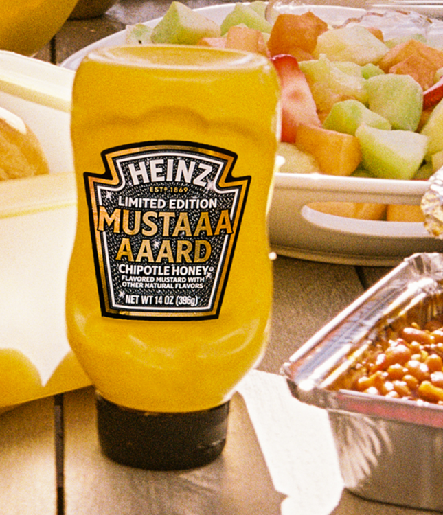 Bright yellow Heinz Limited Edition Mustaaaaard Chipotle Honey bottle, prominently displayed on a wooden picnic table next to a fruit salad, baked beans, and fresh buns, highlighting the perfect summer BBQ condiment.