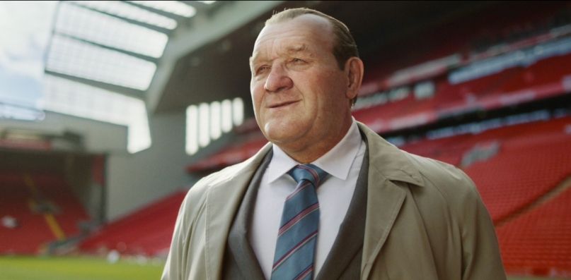 Portrait of computer-generated Bob Paisley, an older man, looking onto a field in a stadium