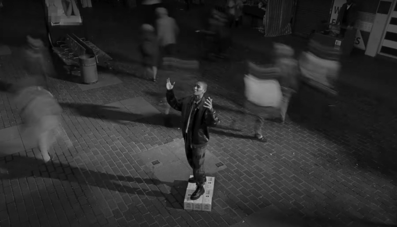 Rapper Roxxxane standing on a soapbox in the middle of a brick sidewalk as people walk by, blurred