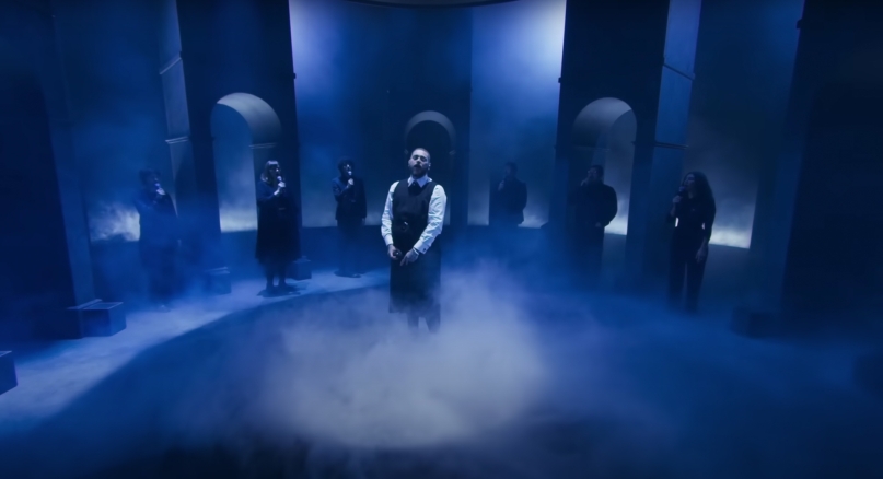 singer Post Malone standing in the middle of a blue room with archways and a semicircle of singers in the background