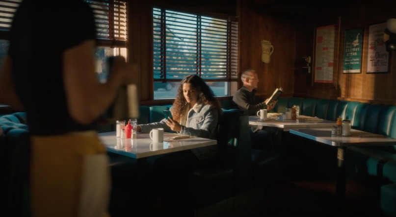 woman sitting in a cafe booth with a patron at the next table behind her and a waitress approaching