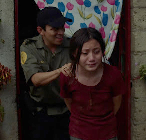 Girl is held by a police officer and they walk out of the house. The girl looks very sad.