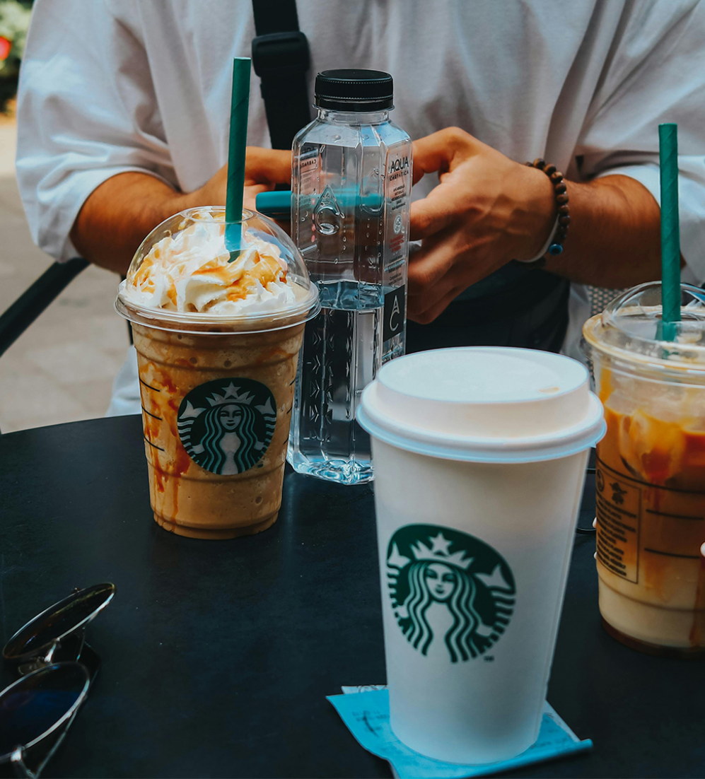 A table outside with all sorts of Starbucks coffe cups on it. At the back of the table you see two hands of a person who is using his mobile.