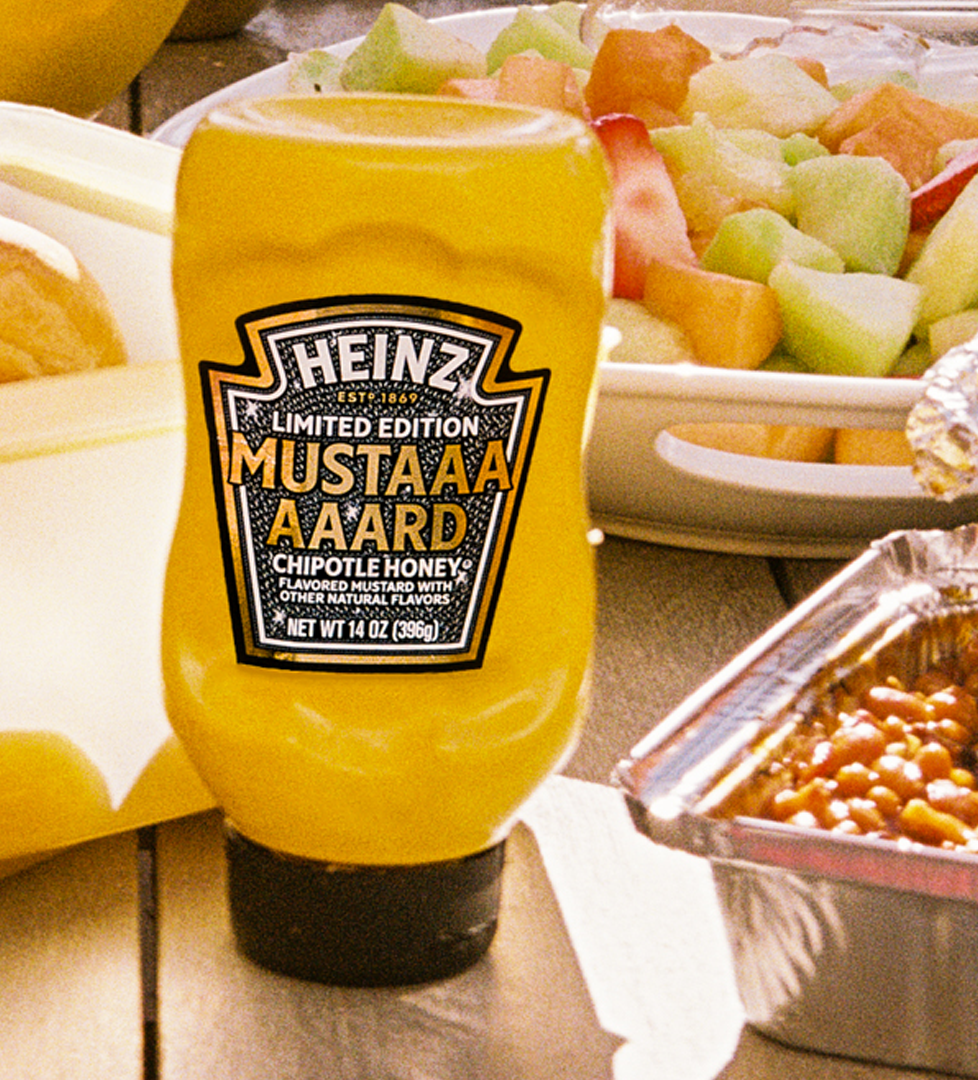 Bright yellow Heinz Limited Edition Mustaaaaard Chipotle Honey bottle, prominently displayed on a wooden picnic table next to a fruit salad, baked beans, and fresh buns, highlighting the perfect summer BBQ condiment.