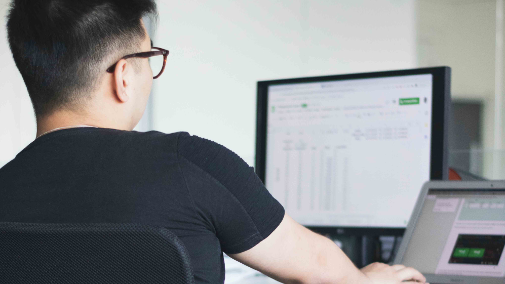 A person staring at a spreadsheet on a desktop and laptop