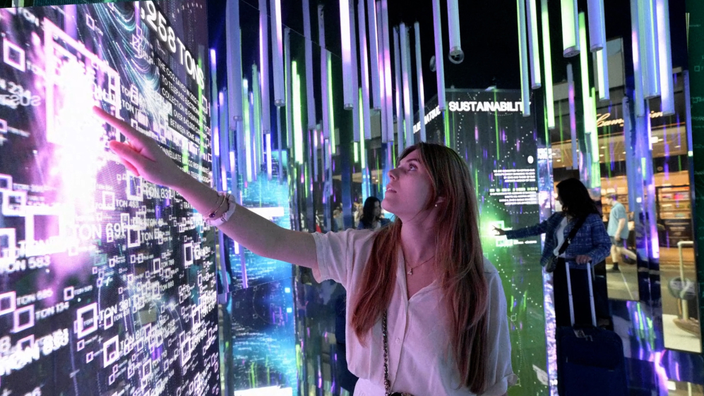 A women standing in front of a touch screen. On the screens there are many lines graphs and letters in different colours.