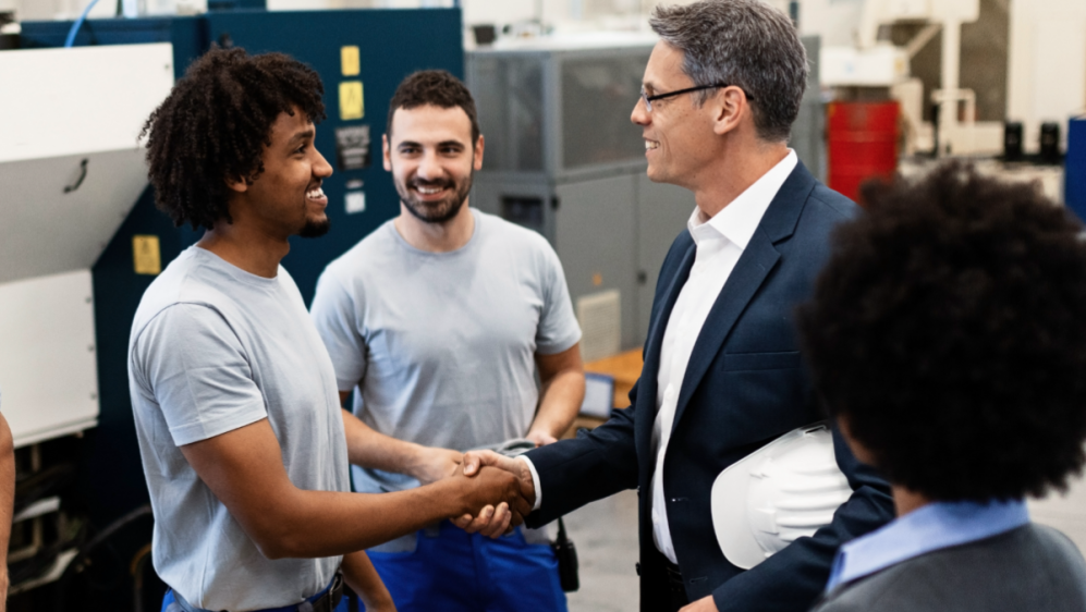 Businessmen shaking hands in a factory. Two factory workers and two people in suits are collaborating.