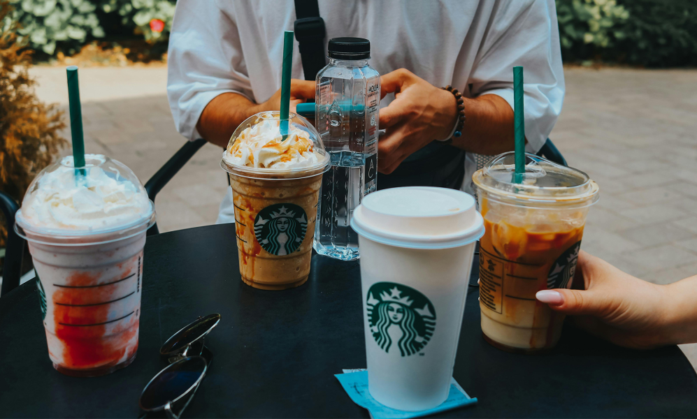 A table outside with all sorts of Starbucks coffe cups on it. At the back of the table you see two hands of a person who is using his mobile.