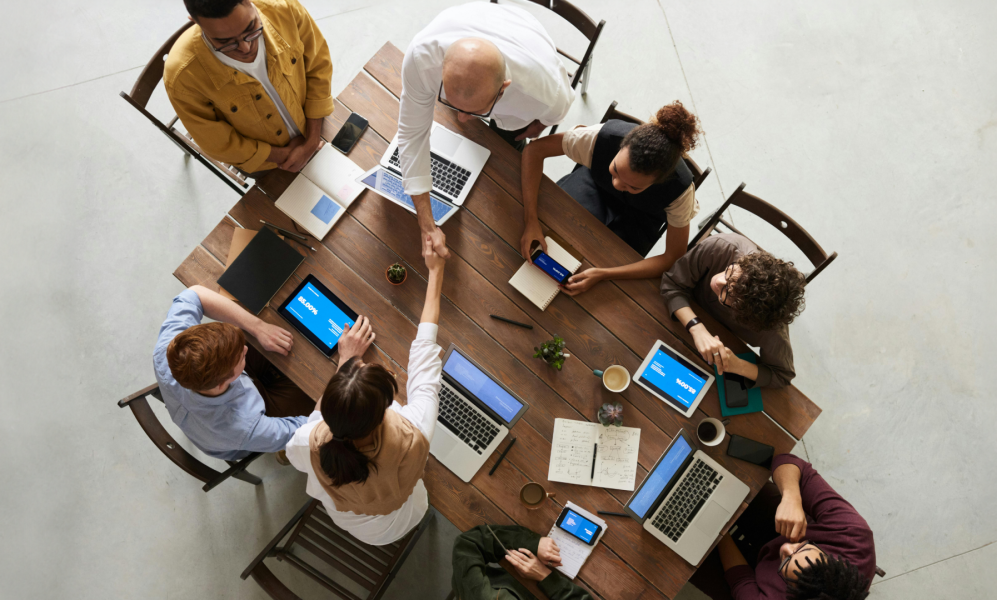 A group of seven people are gathered around a wooden table in a meeting room. They have laptops, tablets, notebooks, and coffee cups in front of them. Two of the individuals are standing and shaking hands over the table, while the others are seated, engaged with their devices or each other. The setting appears collaborative and professional.