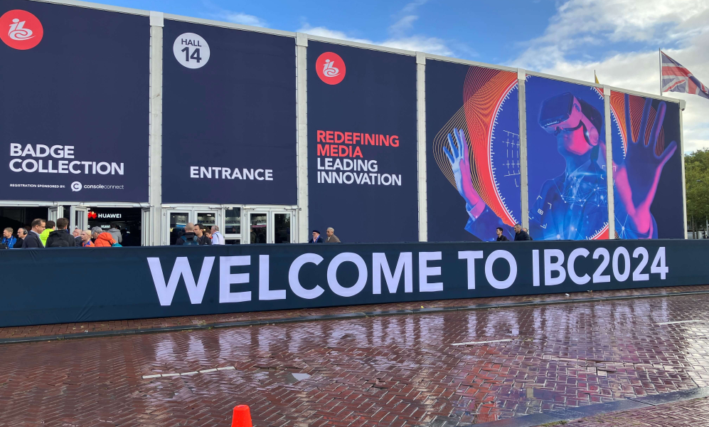 The facade of the IBC convention hall is depicted. Large banners outside display text reading "Badge Collection," "Entrance," and "Redefining Media Leading Innovation." A futuristic image of a person wearing a virtual reality headset is featured. The foreground has a sign that reads "Welcome to IBC2024." People are gathered near the entrance, and a traffic cone is visible on the wet pavement.