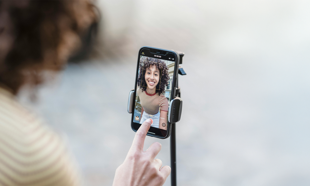 A close-up image of a smartphone mounted on a tripod, capturing a cheerful woman with curly hair wearing a striped shirt during a video recording. A hand is visible in the foreground, about to tap a button on the phone screen.