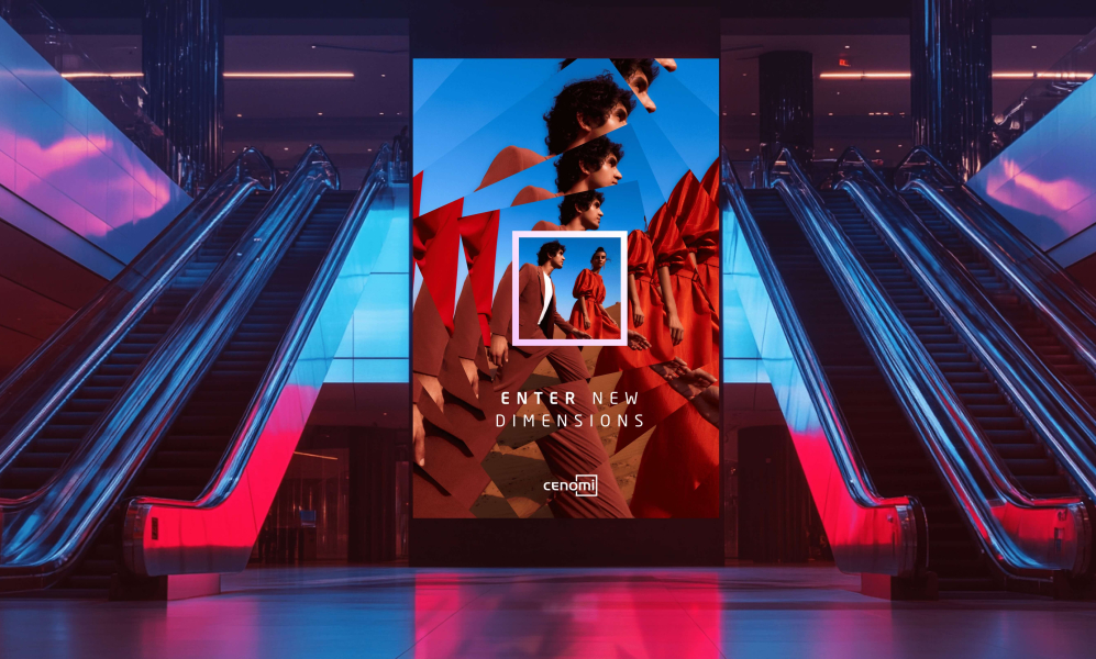 An advertising screen in a transit hub or mall; it is flanked by escalators on each side. The screen reads "Enter new dimensions - Cenomi" and depicts a fashionably dressed man and woman walking in a desert.