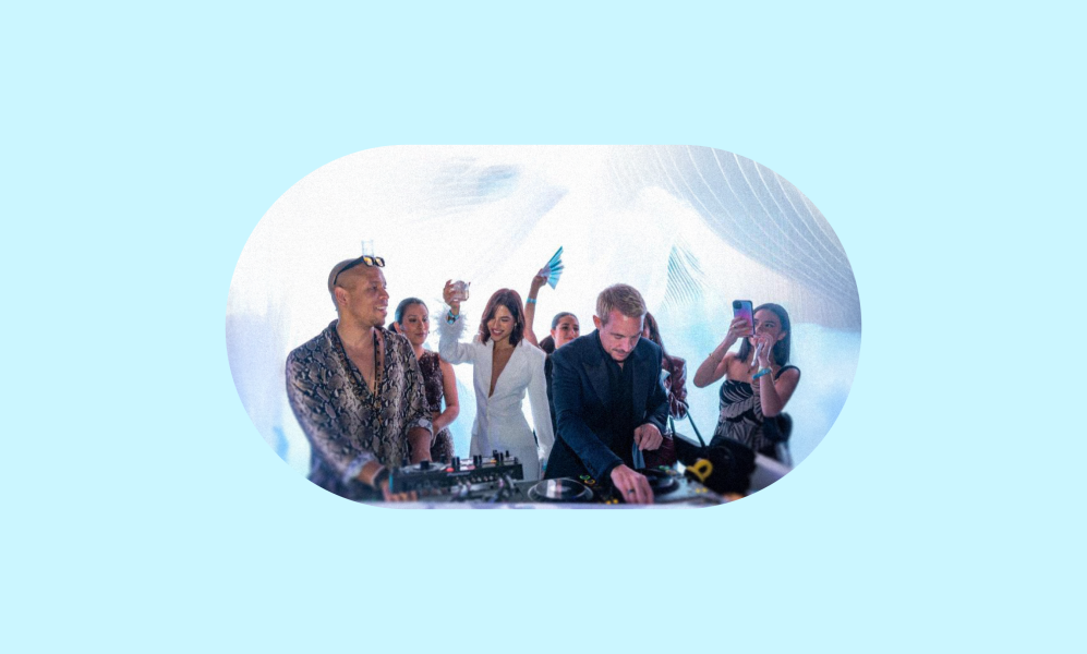 A DJ in a dark suit plays music at a brightly lit event, while another individual in a patterned shirt and sunglasses stands to his left at the DJ booth. Several people are visible behind them, dancing and holding up their phones.