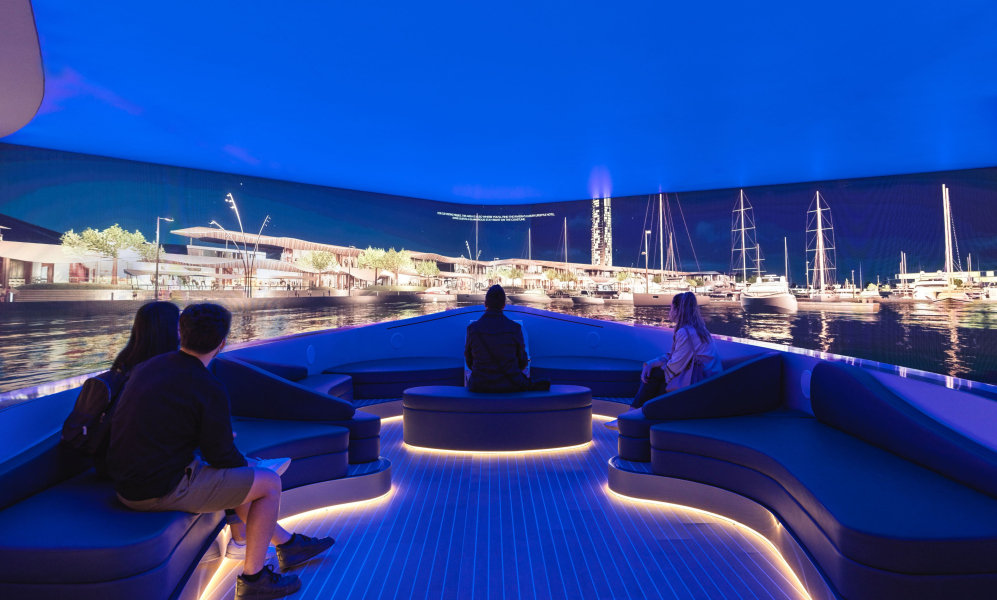 A group of visitors are seated on curved, illuminated benches inside an immersive experience room designed to look like the deck of a yacht. They are watching a panoramic, wrap-around screen displaying a vibrant, photorealistic marina at night.
