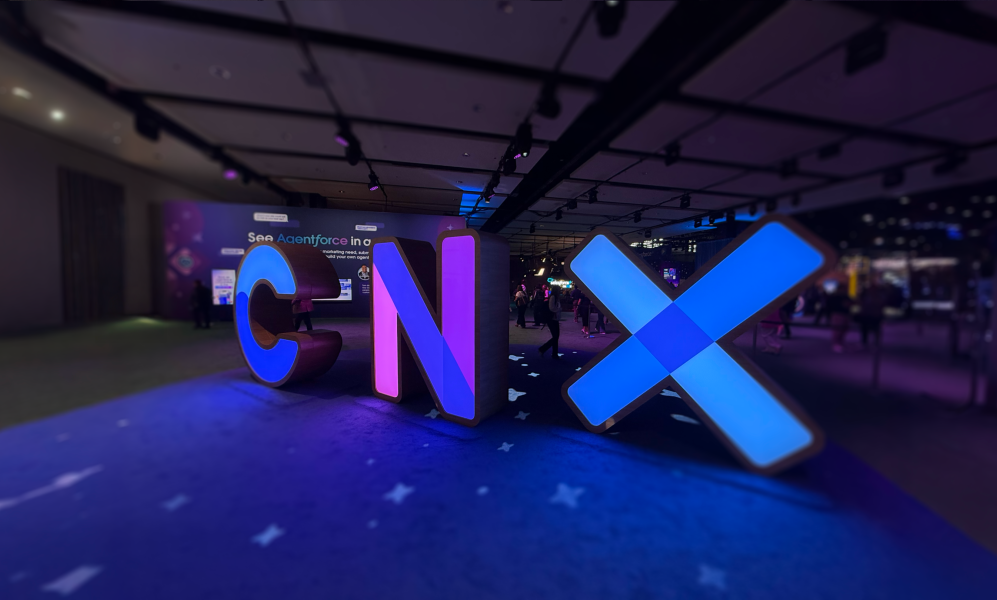 A low-angle, wide shot of large, illuminated, three-dimensional letters spelling "CNX" in a dimly lit event hall. The 'C' glows blue, the 'N' glows purple, and the 'X' glows with a bright blue light. The letters are placed on a dark blue carpet with a subtle pattern, and they cast colorful reflections on the floor. In the blurred background, people can be seen walking around the conference space.