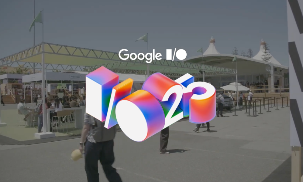 An outdoor shot of the Google I/O 2025 conference on a sunny day. In the foreground is a large, colorful, 3D graphic of the "I/O '25" logo. In the background, attendees walk between large white tents and event structures.