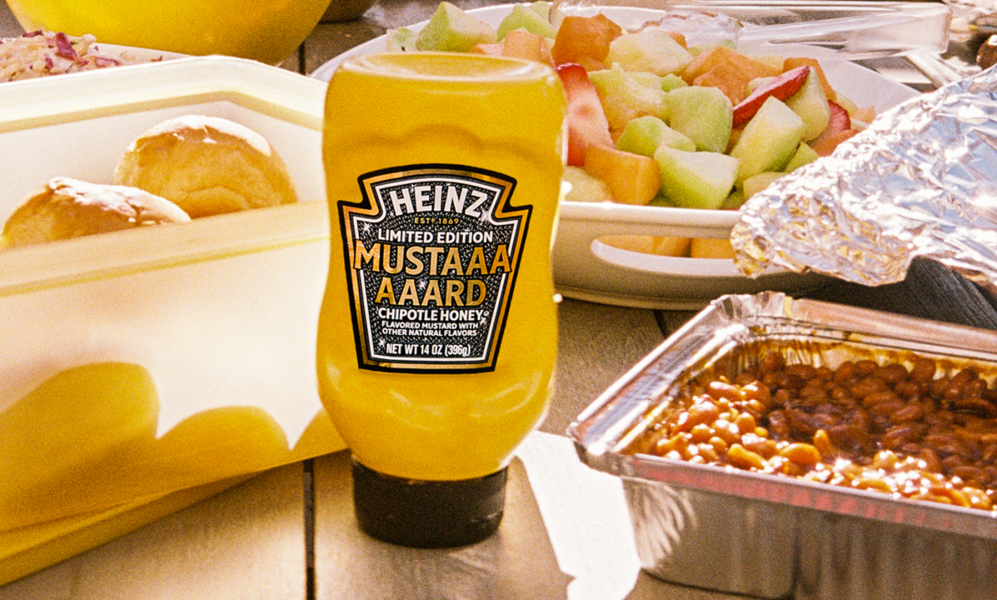 Bright yellow Heinz Limited Edition Mustaaaaard Chipotle Honey bottle, prominently displayed on a wooden picnic table next to a fruit salad, baked beans, and fresh buns, highlighting the perfect summer BBQ condiment.