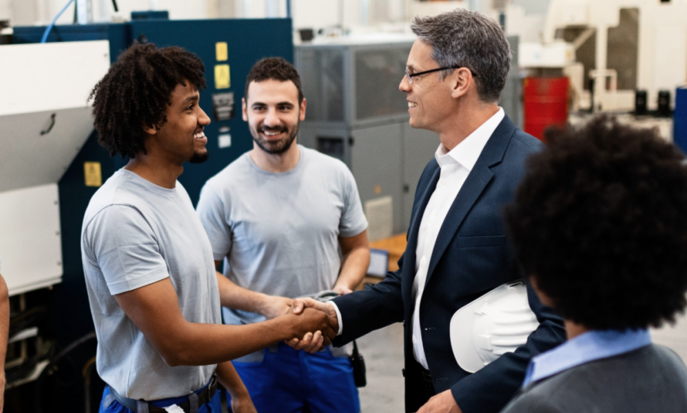 Businessmen shaking hands in a factory. Two factory workers and two people in suits are collaborating.