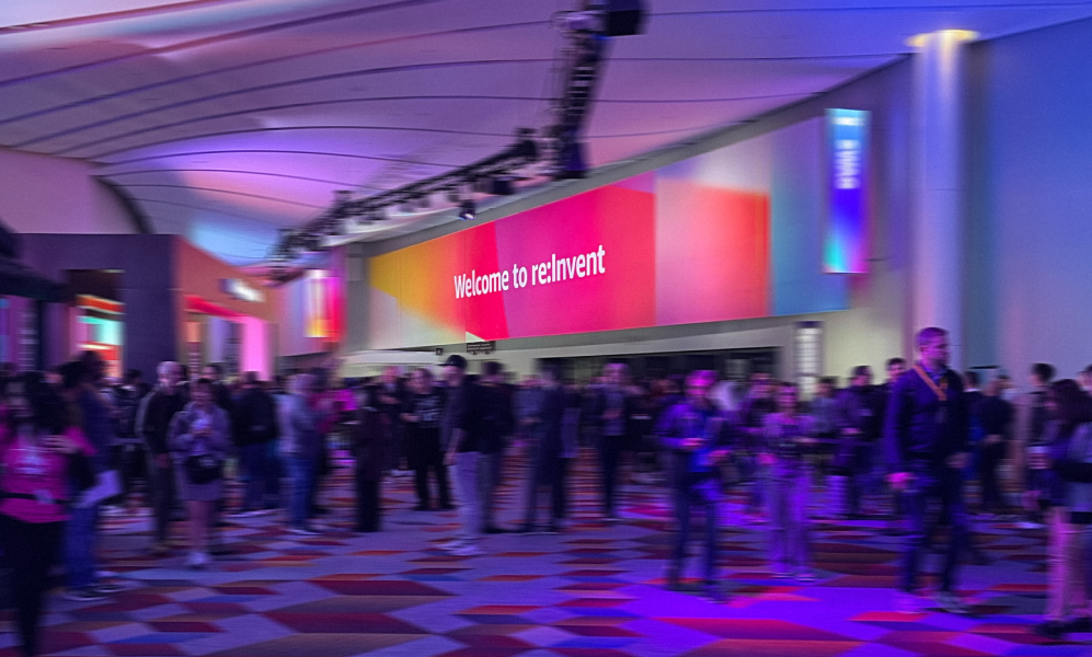 A photograph of a large, crowded convention center hall. A large, curved sign with a colorful pink, orange, and purple gradient background reads "Welcome to re:Invent". The space is illuminated with purple and blue lights, and the floor has a geometric pattern. Numerous attendees are walking around the hall.
