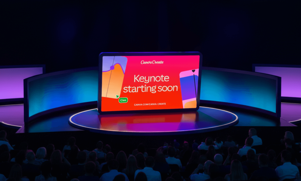 A wide shot of a tech conference stage featuring a large, tablet-shaped central screen that reads "Canva Create" and "Keynote starting soon." The screen is bright red, flanked by curved, glowing blue side panels. In the foreground, a large audience is seated in a darkened theater setting, facing the stage.