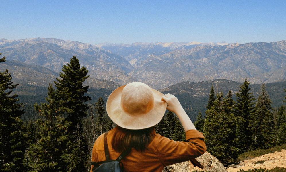 A person seen from behind wearing a wide-brimmed tan hat and an ochre jacket, gazing out at a vast, rugged mountain range and evergreen forest under a clear blue sky.