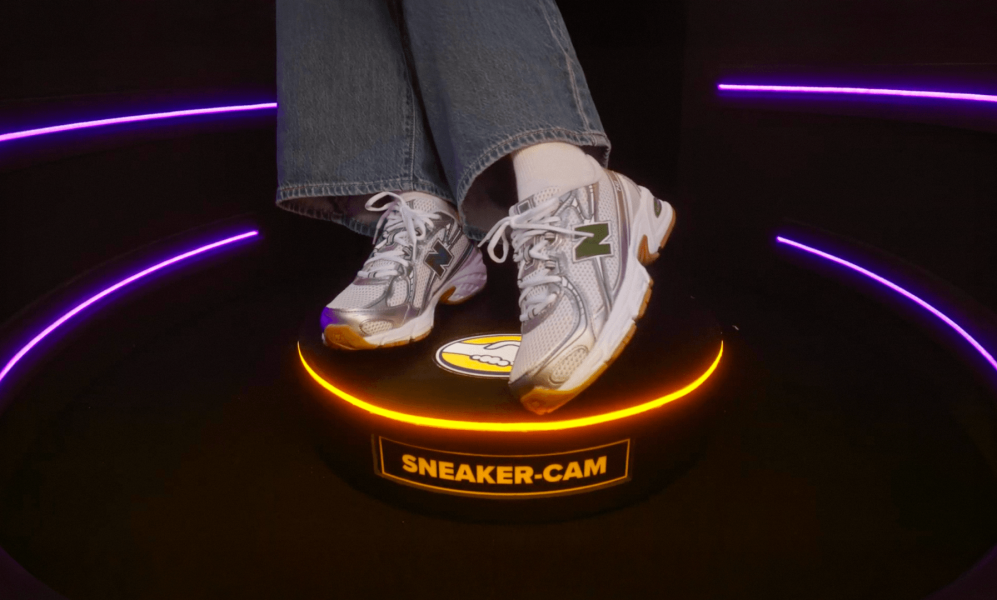 A close-up of a person's feet wearing blue jeans, white socks, and silver and white New Balance sneakers featuring a green "N" logo. They are standing on a small, circular black platform that has a glowing orange ring light around its top edge. The front of the platform displays a yellow label reading "SNEAKER-CAM".
