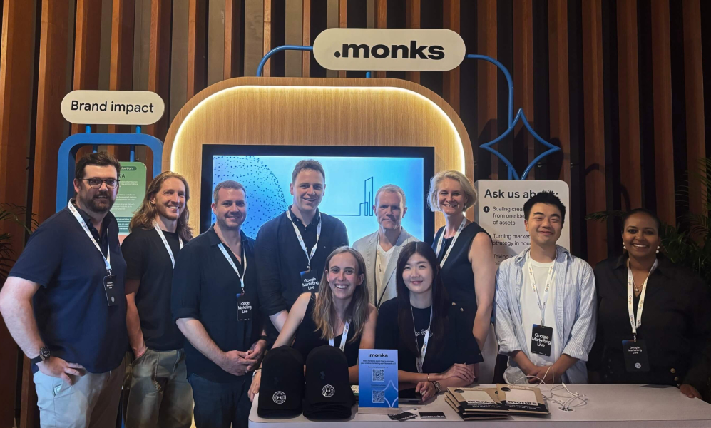 A group of ten smiling professionals posing together at a ".monks" event booth. The attendees are wearing lanyards with "Google Marketing Live" badges. Behind them is a large display screen, an illuminated ".monks" logo, and booth signs reading "Brand impact" and "Ask us about:". In the foreground, a table is set up with branded black caps, brochures, and a small sign featuring a QR code.