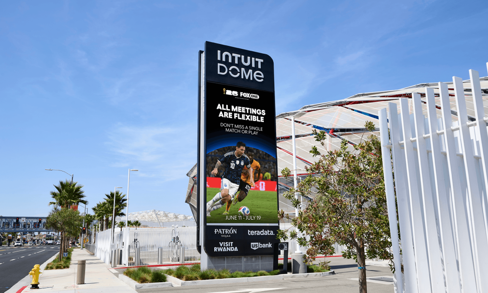 A tall digital billboard outside the Intuit Dome displaying a FIFA World Cup advertisement featuring soccer player Lionel Messi in action. The screen reads "ALL MEETINGS ARE FLEXIBLE" above the dates June 11 through July 19, alongside logos for Fox and various corporate sponsors like Patron and US Bank. The sign stands on a sunny, palm-tree-lined sidewalk with the stadium's exterior visible in the background behind a white fence.