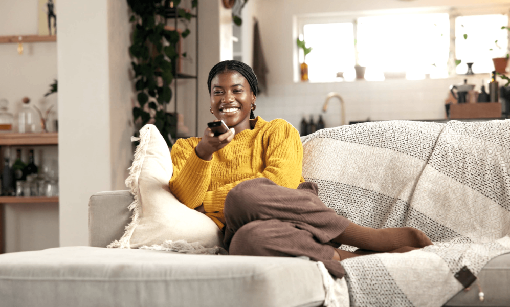 A smiling woman in a yellow sweater relaxes on a couch, holding a TV remote and looking toward the camera.