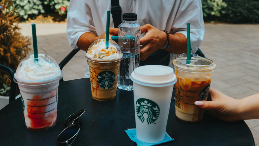 A table outside with all sorts of Starbucks coffe cups on it. At the back of the table you see two hands of a person who is using his mobile.