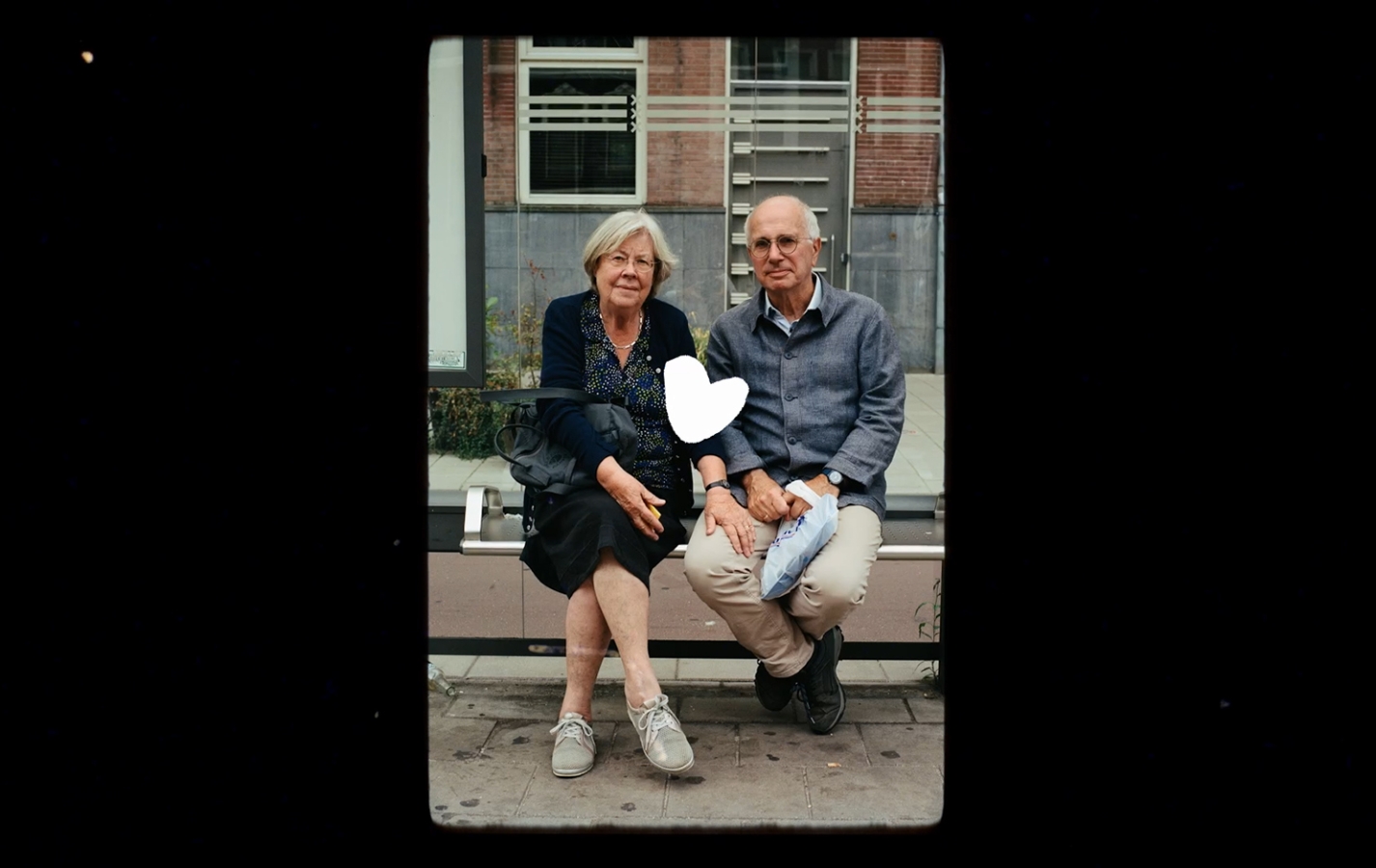 An elderly couple sitting in a waiting booth of the public transport. There is an illustrated white heart pleaced between them.