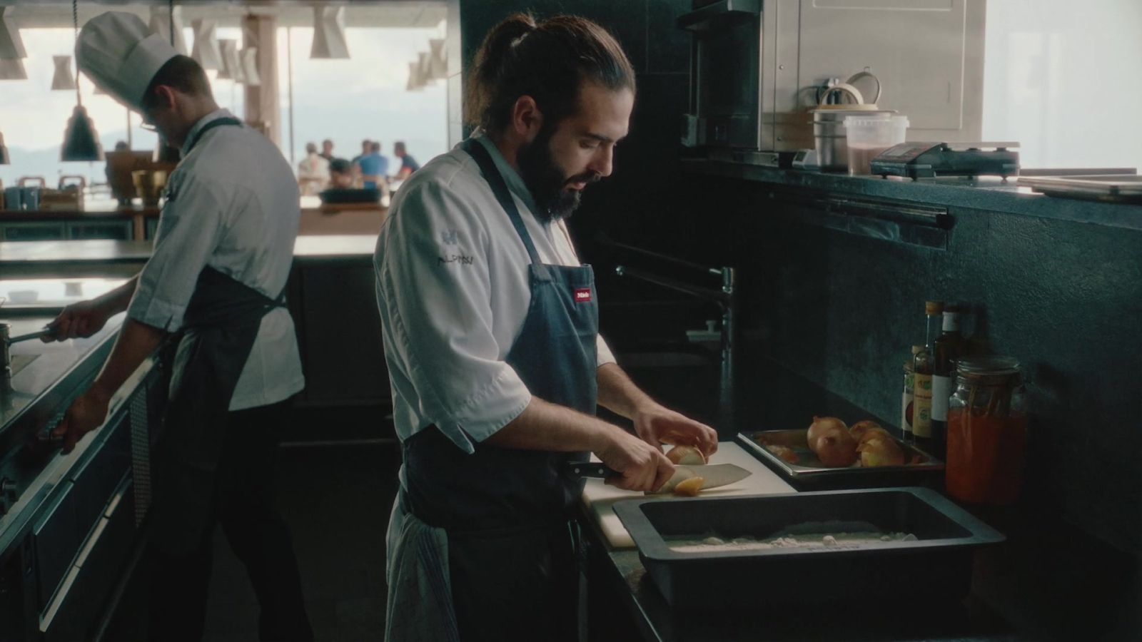 A sous-chef in a kitchen cutting an onion. He wears and apron with Miele logo on it.