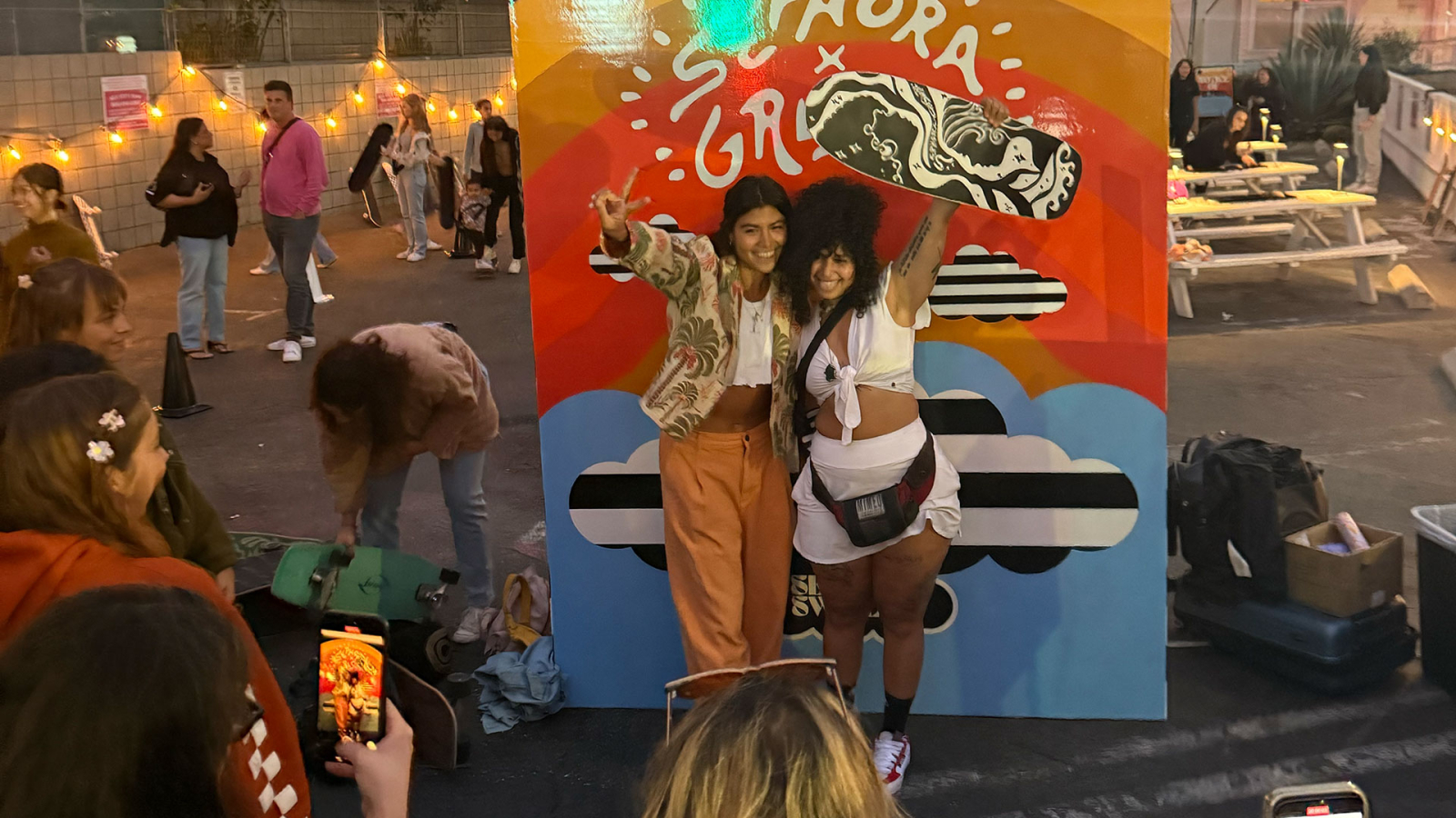 The photo captures a lively event scene where two women pose happily in front of a colorful backdrop that features the Sephora logo, surrounded by festive string lights. In the foreground, a group of people is engaged in conversation, adding to the cheerful atmosphere.