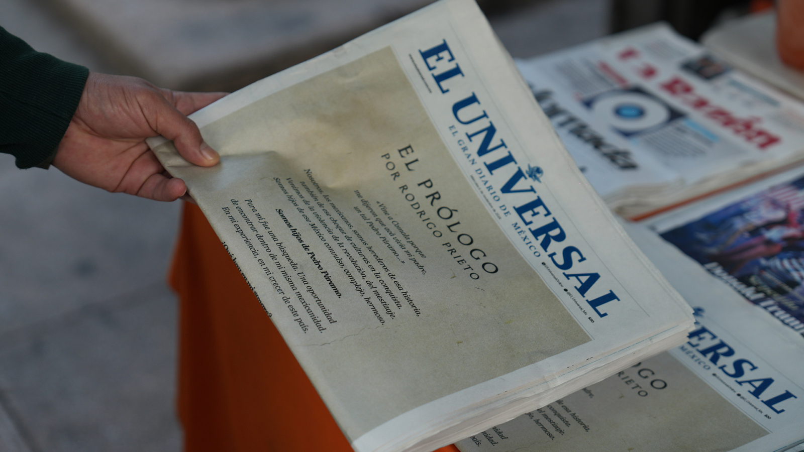 The image shows a hand holding a newspaper titled "El Universal," featuring an article titled "El Prólogo" by Rodrigo Prieto, centered on a table with more newspapers in the background.