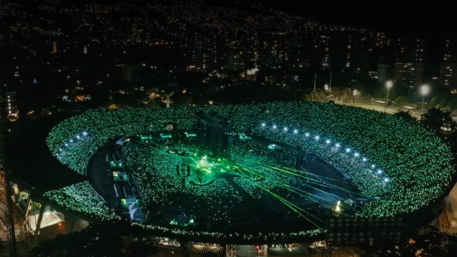 The image shows a large outdoor venue illuminated with green lights, featuring an audience with laser beams, set against a cityscape at night.