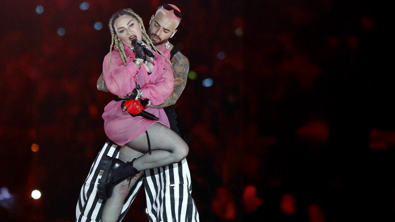 The image shows a singer dressed in pink and fishnet stockings performing passionately while being embraced from behind on a dark stage with blurred lights in the background.
