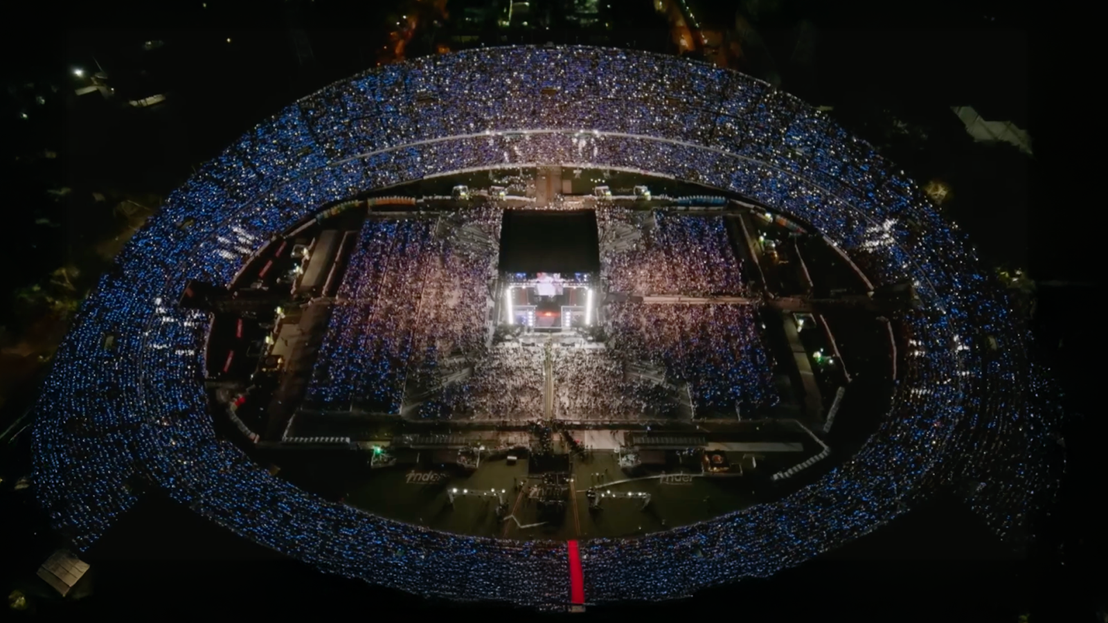 The image shows an aerial view of a large, illuminated stadium filled with thousands of people at night, with bright lights outlining the structure.